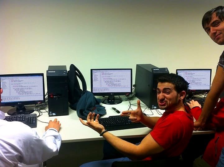 Ein Computerlabor an der Uni: Nikos dreht sich vor einer Reihe von Monitoren mit Code zur Kamera.